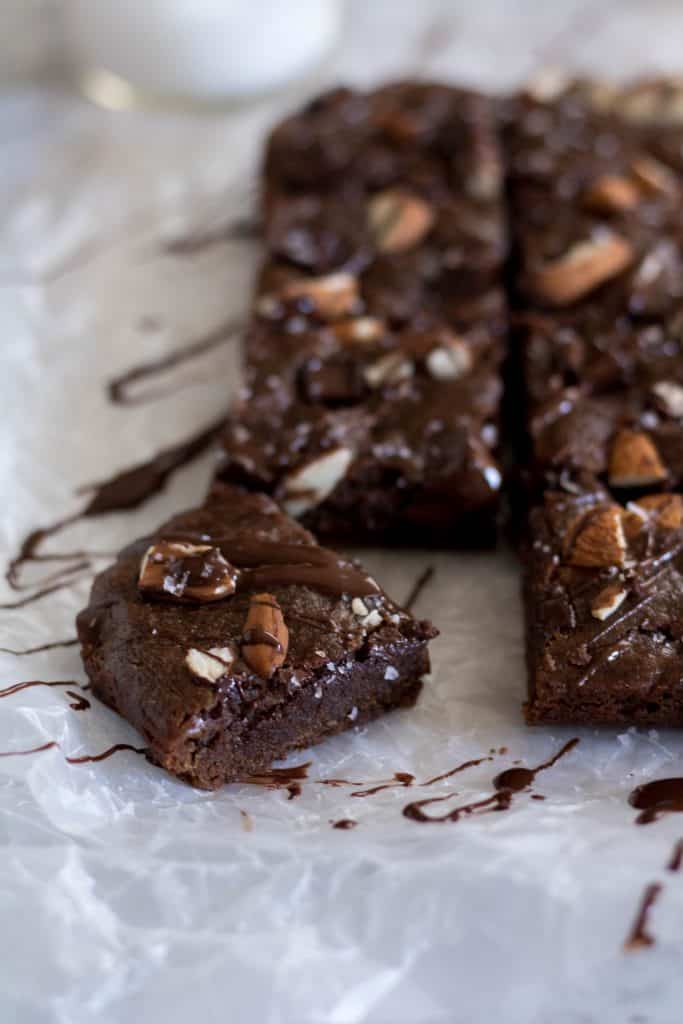 Fudgy vegan double chocolate brownies sliced into squares on parchment paper topped with chopped almonds and a chocolate drizzle with one piece pulled away