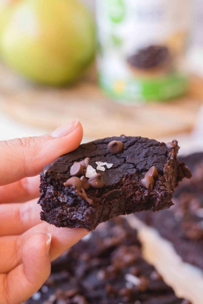 Hand holding a vegan black bean brownie with chocolate chips and flaky sea salt on top showing the dense fudgy interior with an avocado in the background