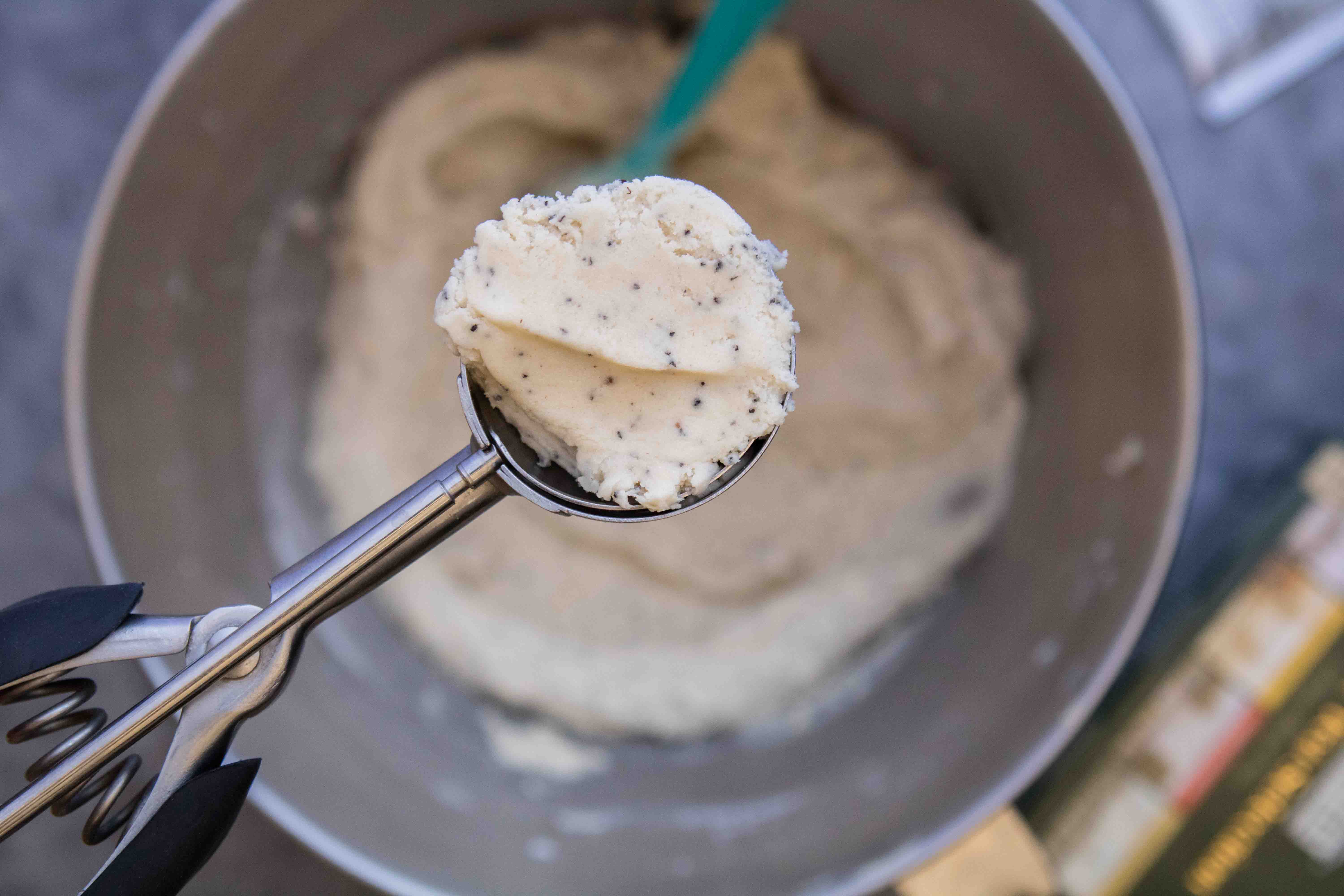 A cookie scoop lifting Earl Grey cookie dough speckled with tea leaves out of a mixing bowl.