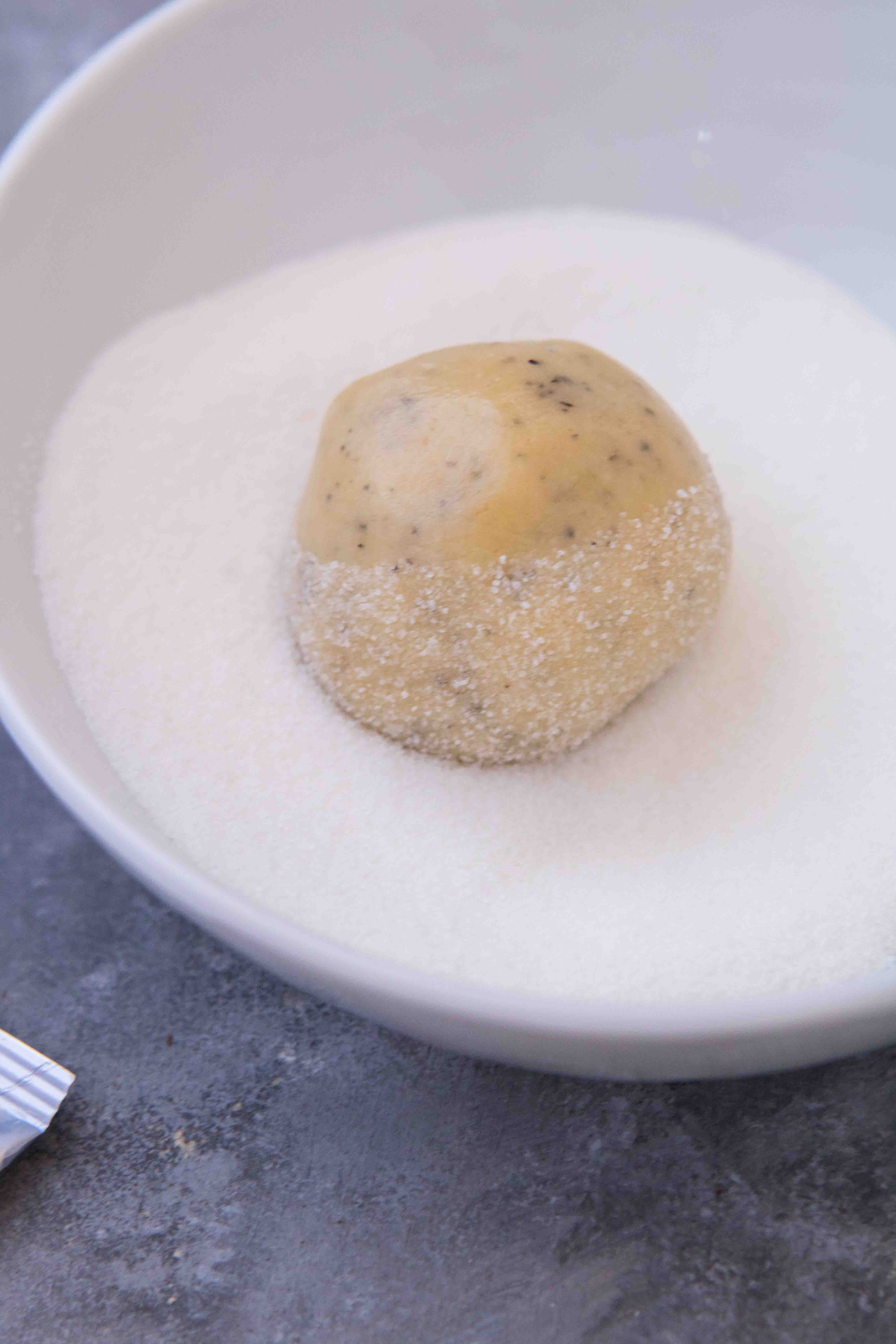 An Earl Grey cookie dough ball being rolled in a white bowl of granulated sugar.