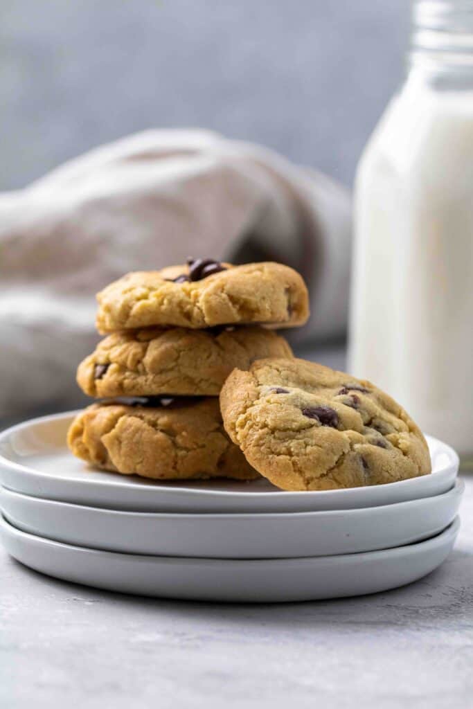 Soft and chewy air fryer chocolate chip cookies stacked on white plates with a glass bottle of milk in the background