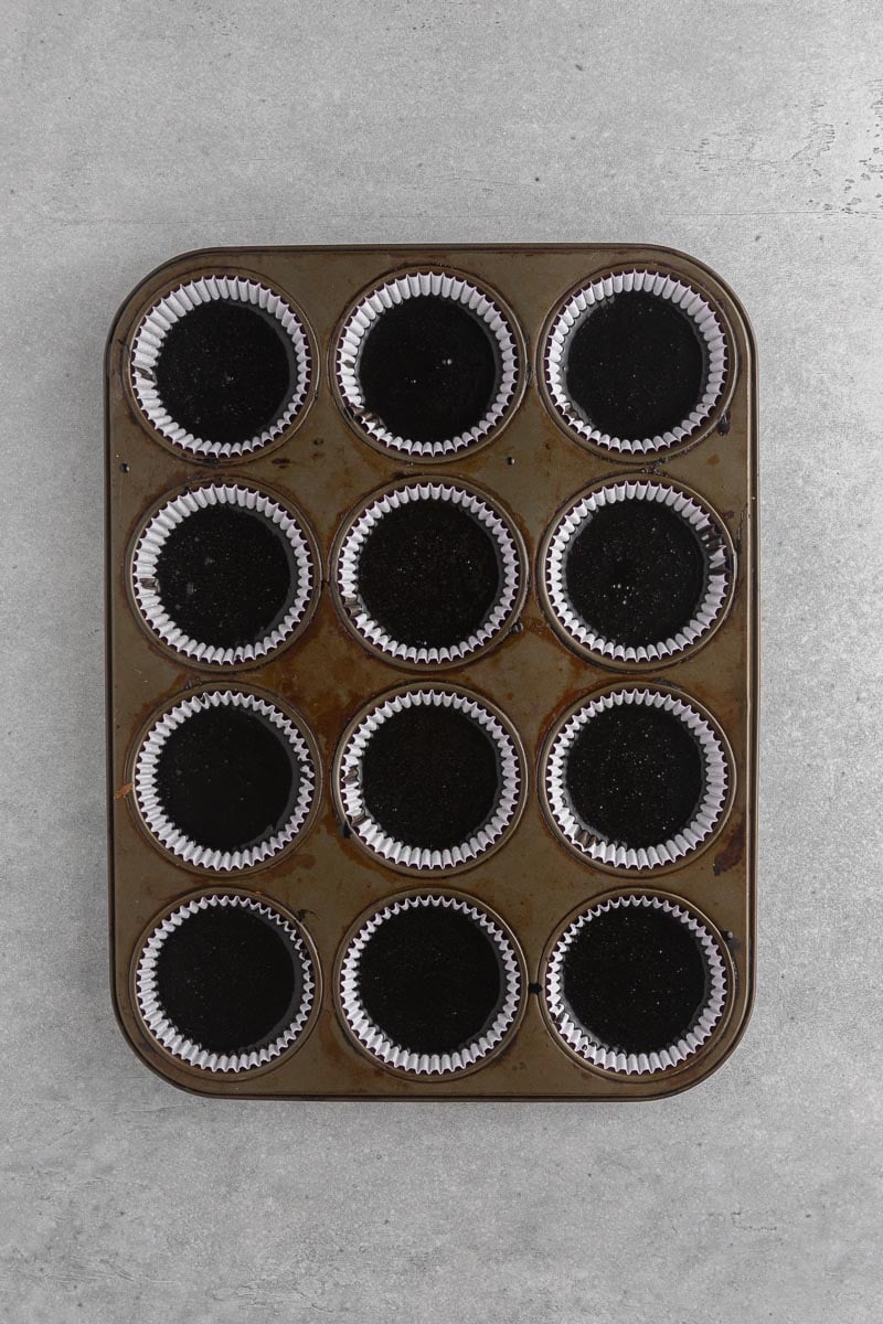 Black velvet cupcake batter divided between paper liners in a 12-cup muffin tin.