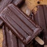 Homemade fudgesicles on wooden sticks piled on parchment paper with chocolate chips scattered around.