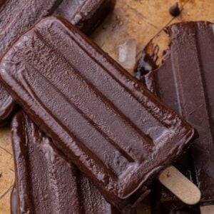 Homemade fudgesicles on wooden sticks piled on parchment paper with chocolate chips scattered around.