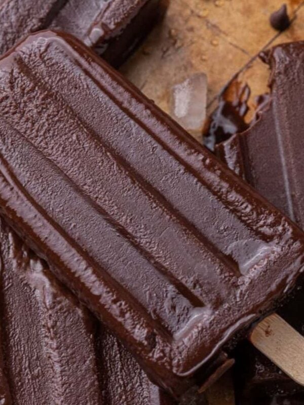 Homemade fudgesicles on wooden sticks piled on parchment paper with chocolate chips scattered around.