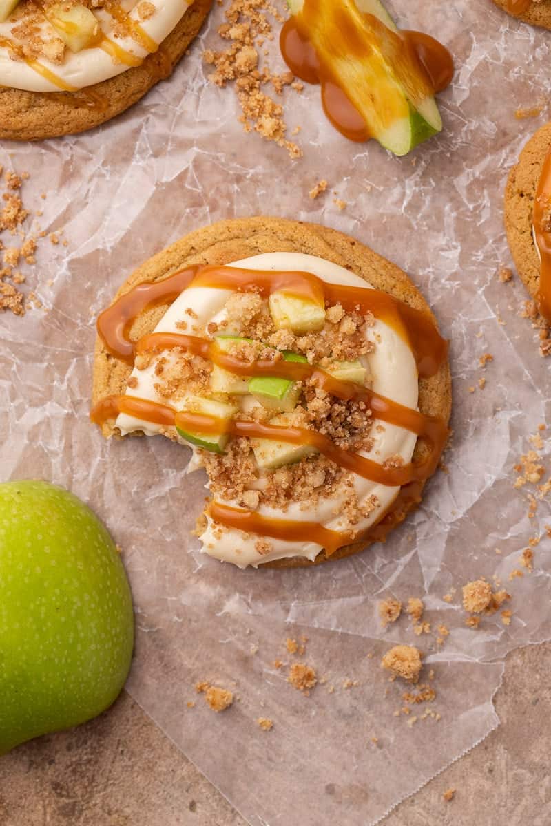 Overhead view of a caramel apple cookie with a bite taken out, topped with cream cheese frosting, diced green apple, streusel, and a caramel drizzle.