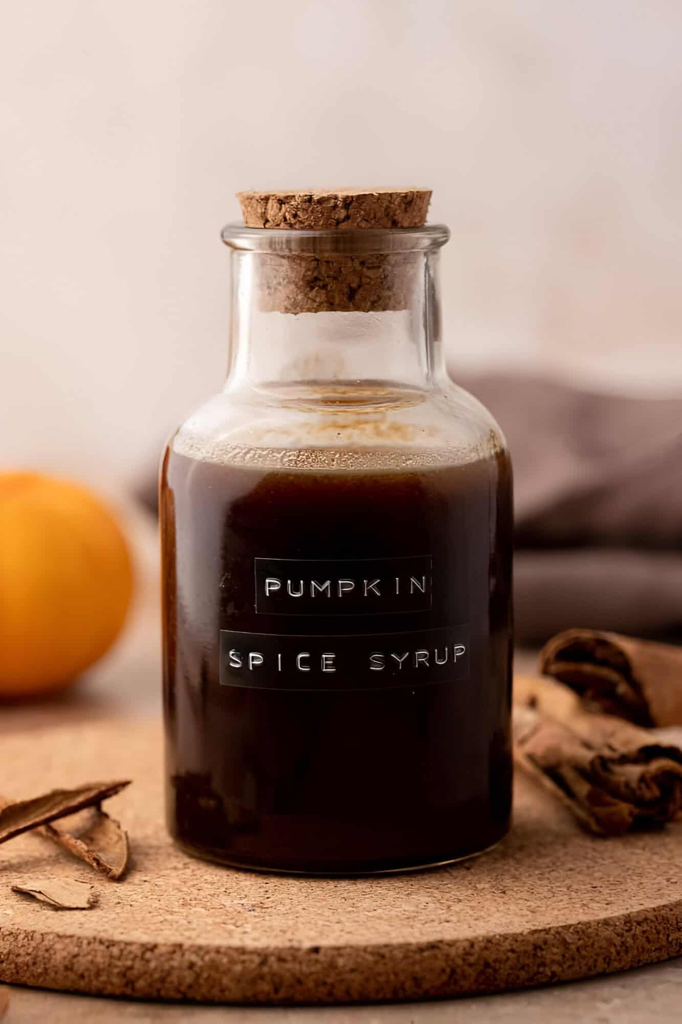 Homemade Starbucks pumpkin spice syrup in a glass bottle beside a cinnamon stick and coffee.