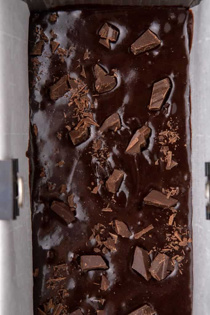 Glossy brownie batter topped with chocolate chunks in a parchment lined loaf pan.