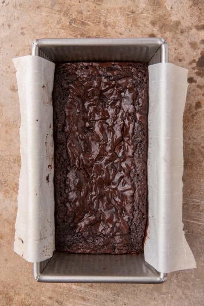 Baked brownies cooling in a parchment lined loaf pan with a deeply crackled top.