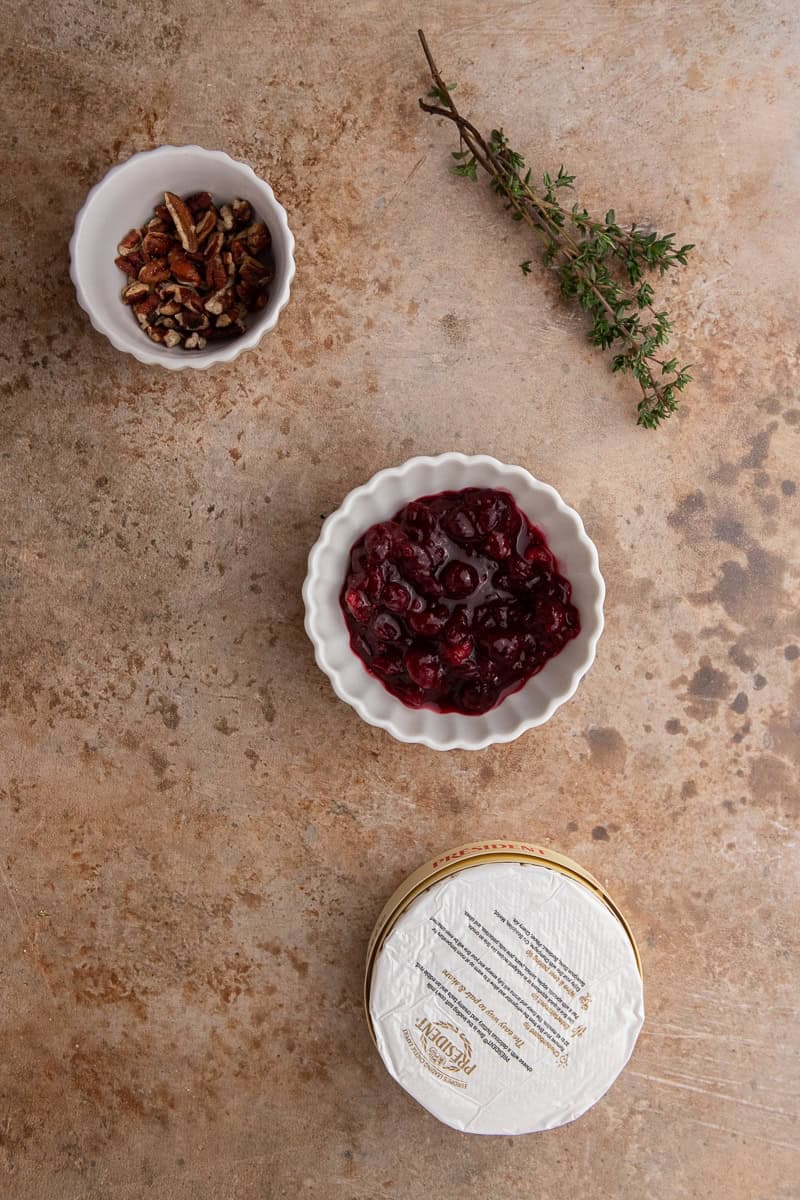 Small bowls of toasted chopped pecans and cranberry sauce next to a wheel of brie and a sprig of fresh thyme.