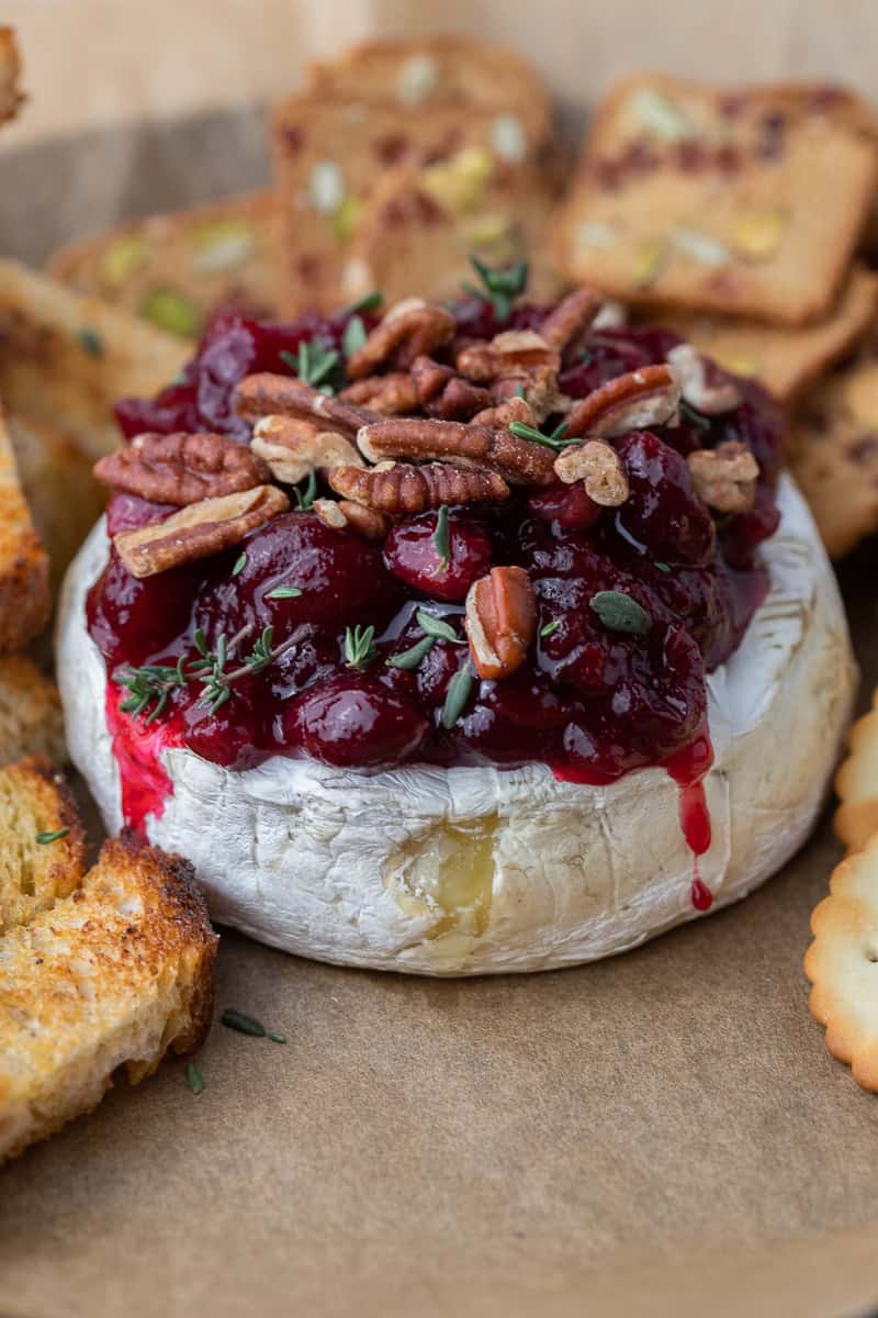 Warm baked brie topped with cranberry sauce, chopped pecans, and thyme, with sauce dripping down the sides.