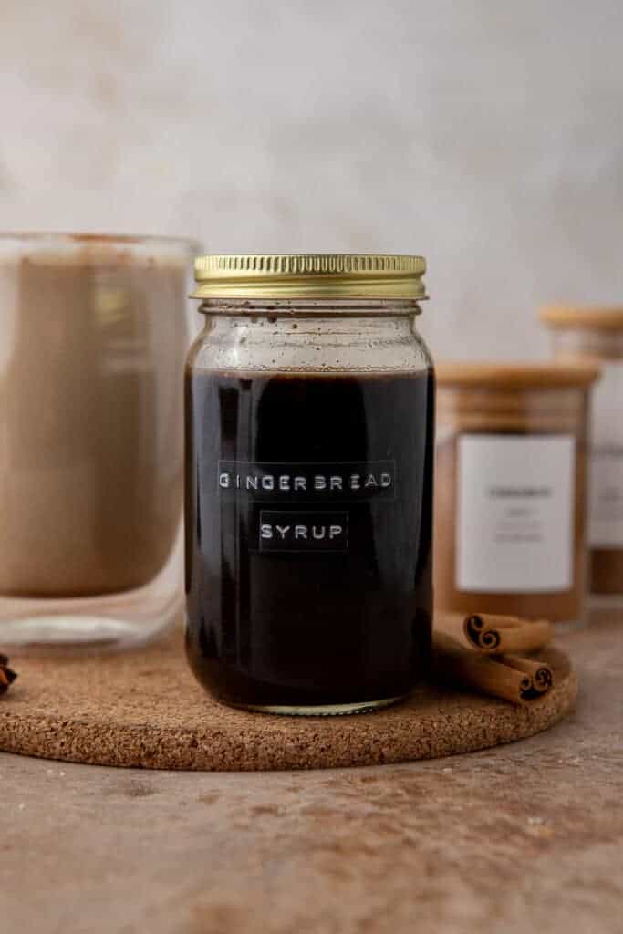 A styled photo of gingerbread syrup in a jar with festive background elements like cinnamon sticks and a latte to show serving inspiration.