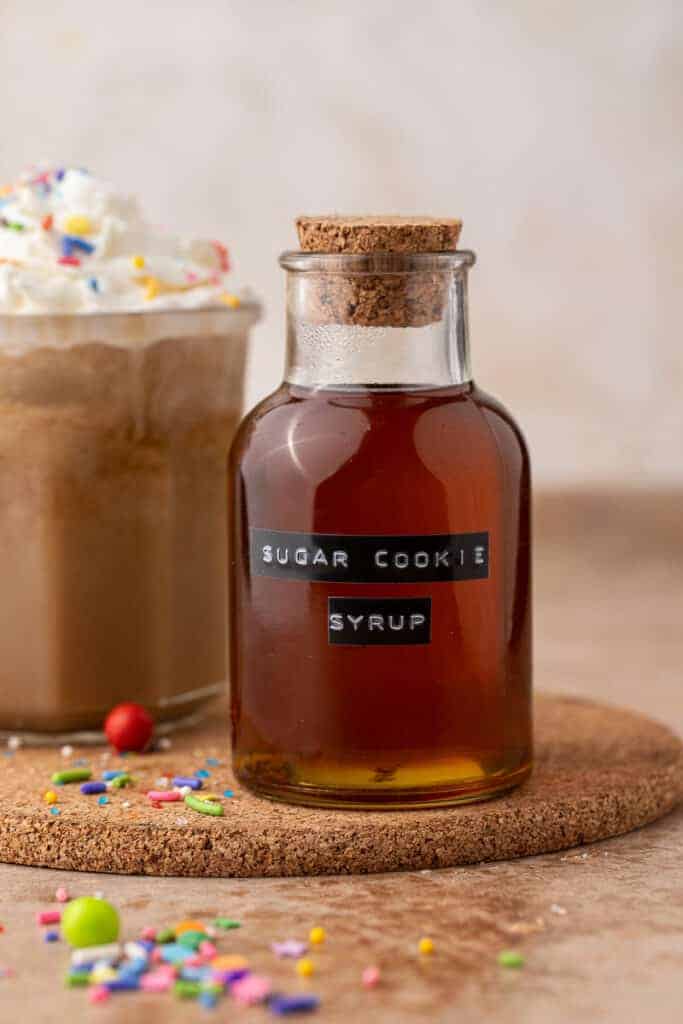 Homemade Starbucks sugar cookie syrup in a corked glass bottle with a holiday sugar cookie latte behind it