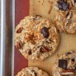 Overhead view of baked cowboy cookies with chocolate chunks, pecans and toasted coconut on a lined baking sheet.