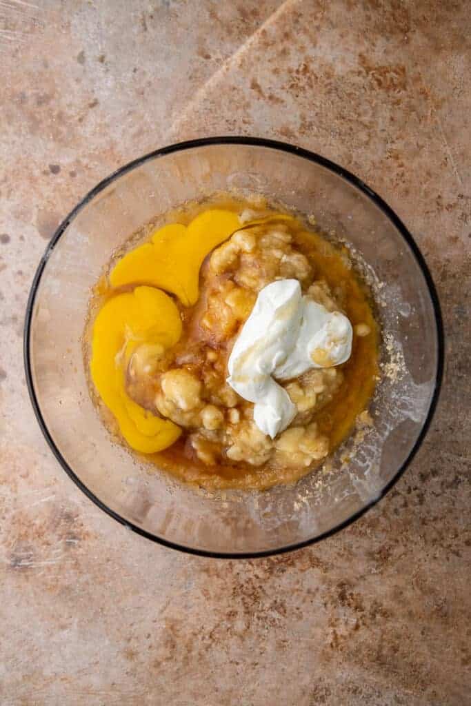 Wet ingredients in a glass mixing bowl including egg yolks, mashed ripe bananas, and Greek yogurt before mixing