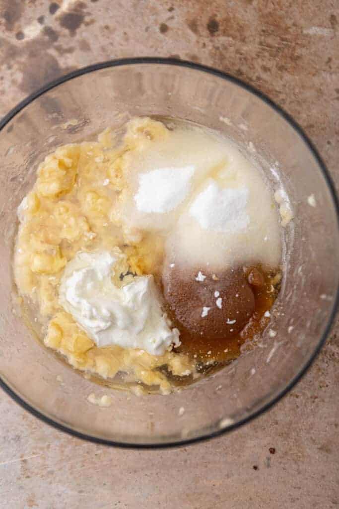 Mashed bananas with sour cream, sugar, and oil in a glass mixing bowl for eggless banana bread