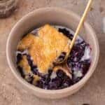 A serving of blueberry cobbler in a bowl with a gold spoon.