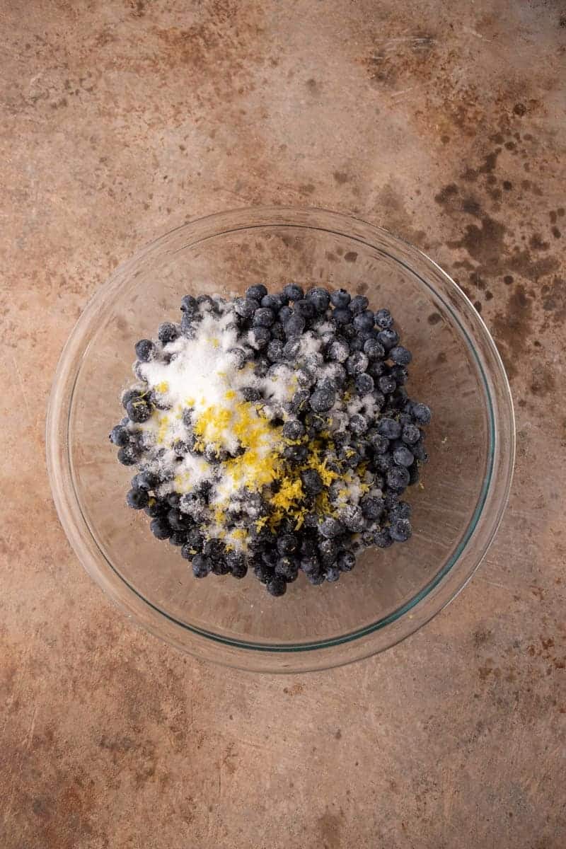 Fresh blueberries tossed with sugar and lemon zest in a glass bowl.