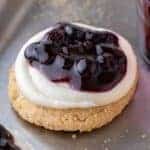 A blueberry cheesecake cookie topped with cream cheese frosting and glossy blueberry topping on a baking sheet sprinkled with graham crumbs.