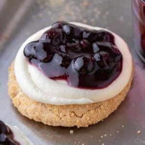 A blueberry cheesecake cookie topped with cream cheese frosting and glossy blueberry topping on a baking sheet sprinkled with graham crumbs.