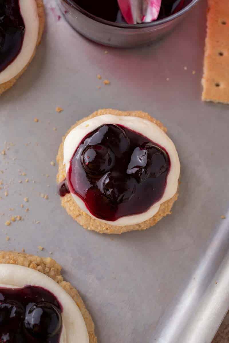 Overhead view of a cookie topped with cream cheese frosting and blueberry topping on a baking sheet.