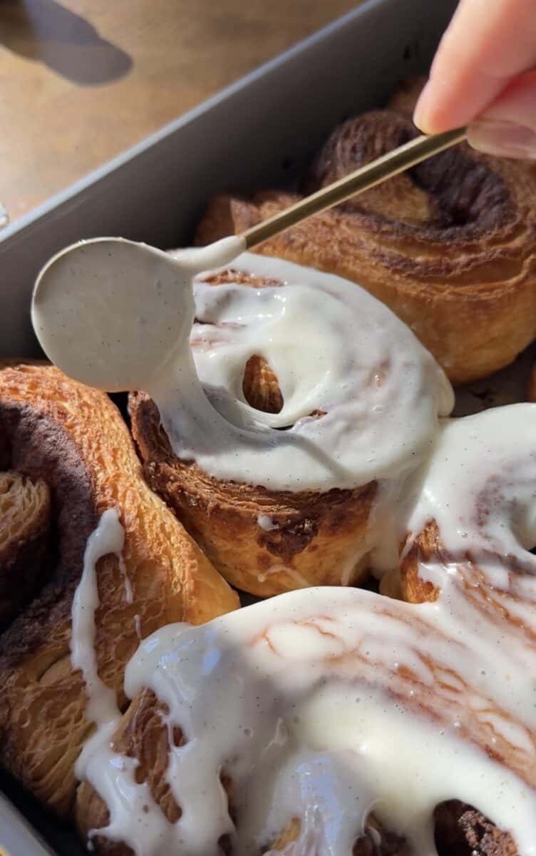 Cream cheese glaze being spooned over warm croissant cinnamon rolls in a pan