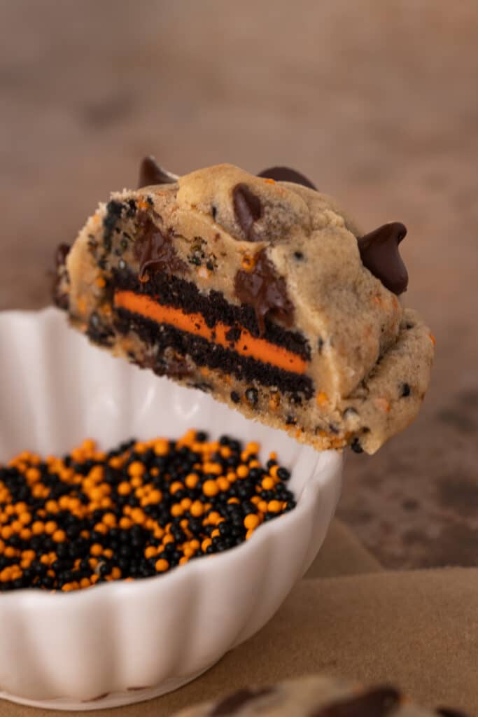 a split Halloween Oreo stuffed chocolate chip cookie on top of a bowl of sprinkles.
