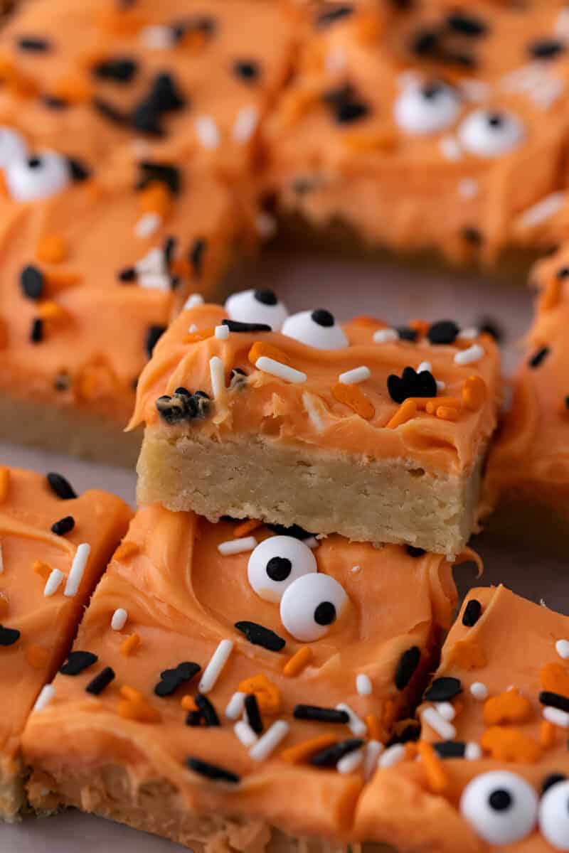 Halloween sugar cookie bars with orange frosting, candy eyeballs, and spooky sprinkles, one bar stacked on top.
