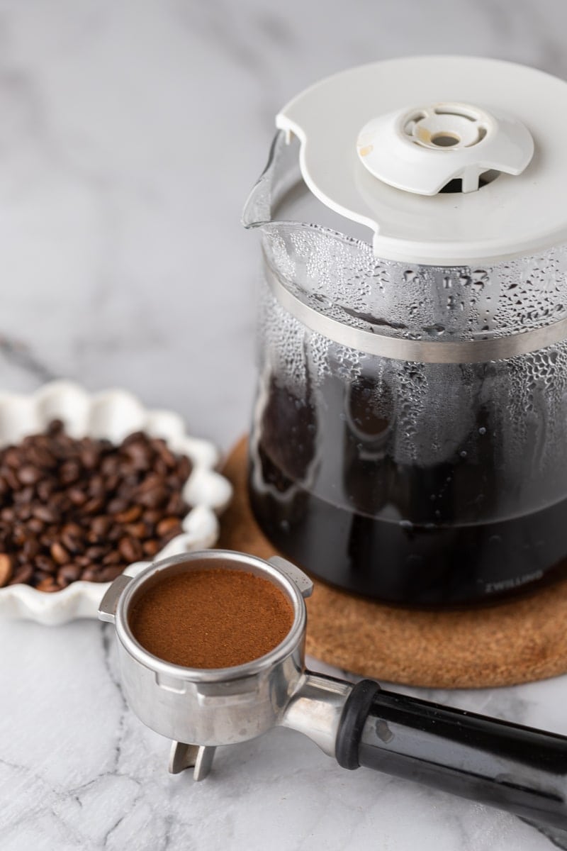A pot of freshly brewed coffee on a cork coaster next to a portafilter of ground espresso and a bowl of coffee beans.