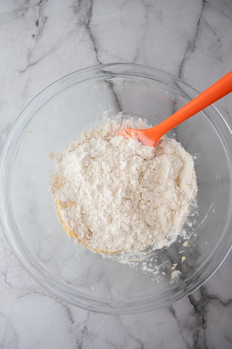 Heat-treated flour added to the lemon butter mixture in a glass bowl with an orange spatula.
