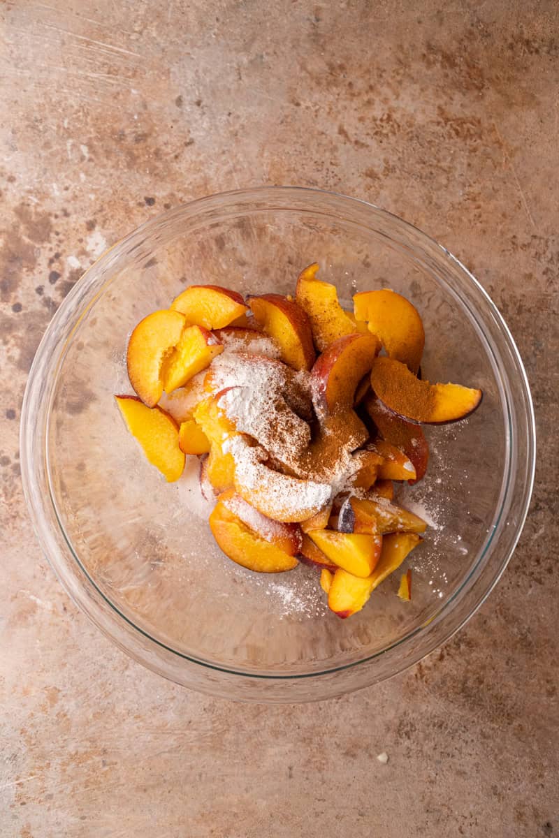 Sliced peaches tossed with sugar and spices in a mixing bowl.