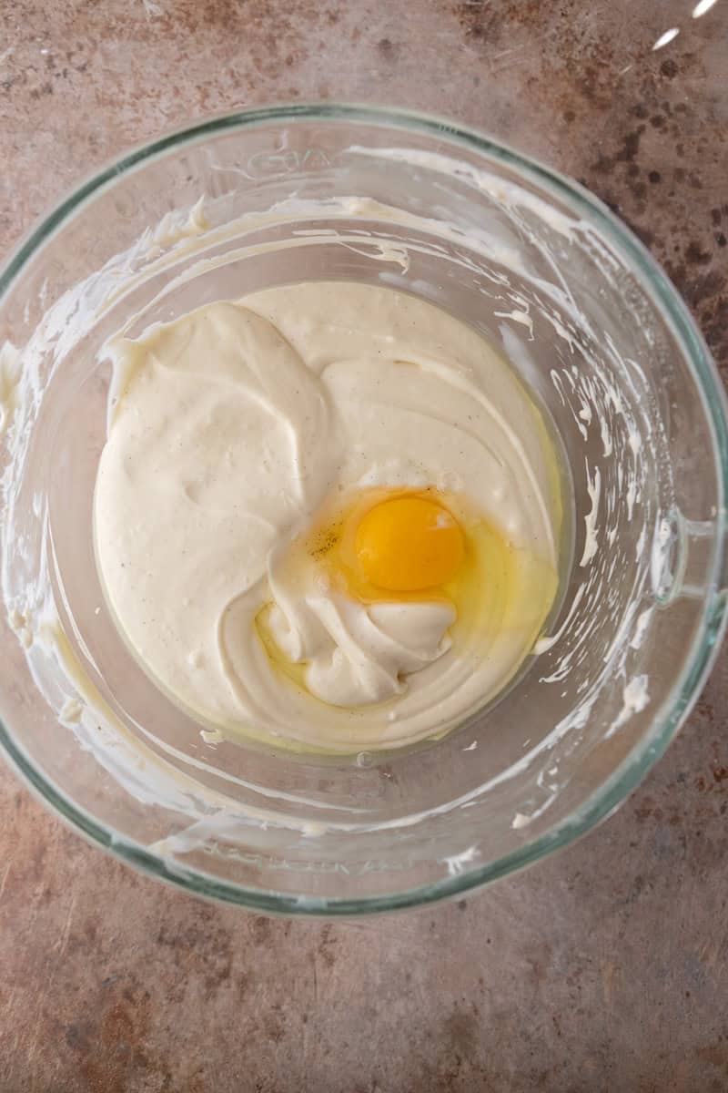An egg being added to the apple crisp cheesecake filling in a glass bowl.