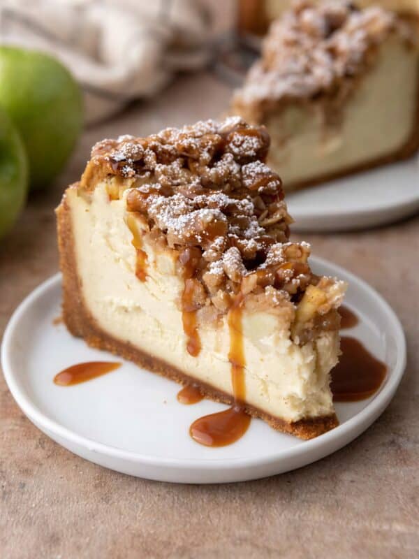 A thick slice of apple crisp cheesecake with oat streusel, diced apples, and caramel drizzle on a white plate.