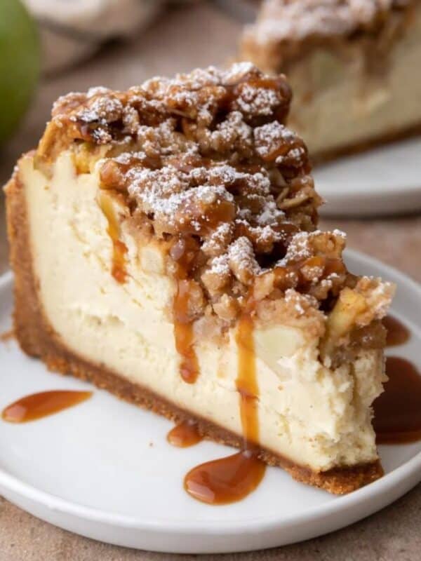 A thick slice of apple crisp cheesecake with oat streusel, diced apples, and caramel drizzle on a white plate.