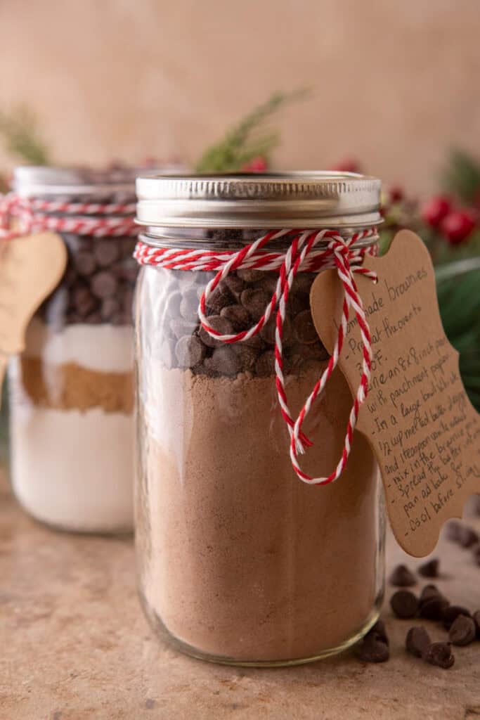 Two mason jars filled with layered homemade brownie mix dry ingredients tied with red and white baker's twine and handwritten recipe tags with holiday greenery in the background