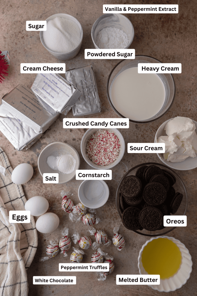 Overhead view of cheesecake ingredients including cream cheese, Oreos, sour cream, heavy cream, extracts, crushed candy canes, and Lindt peppermint truffles.