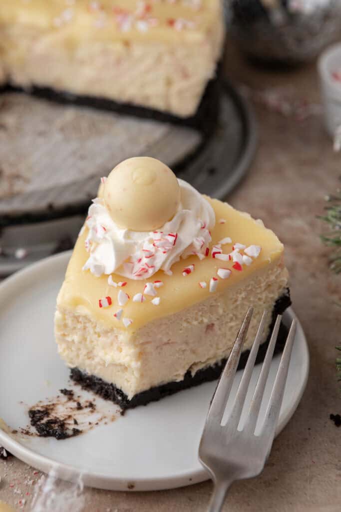 Slice of peppermint cheesecake on a white plate with whipped cream, candy cane pieces, and truffle on top.