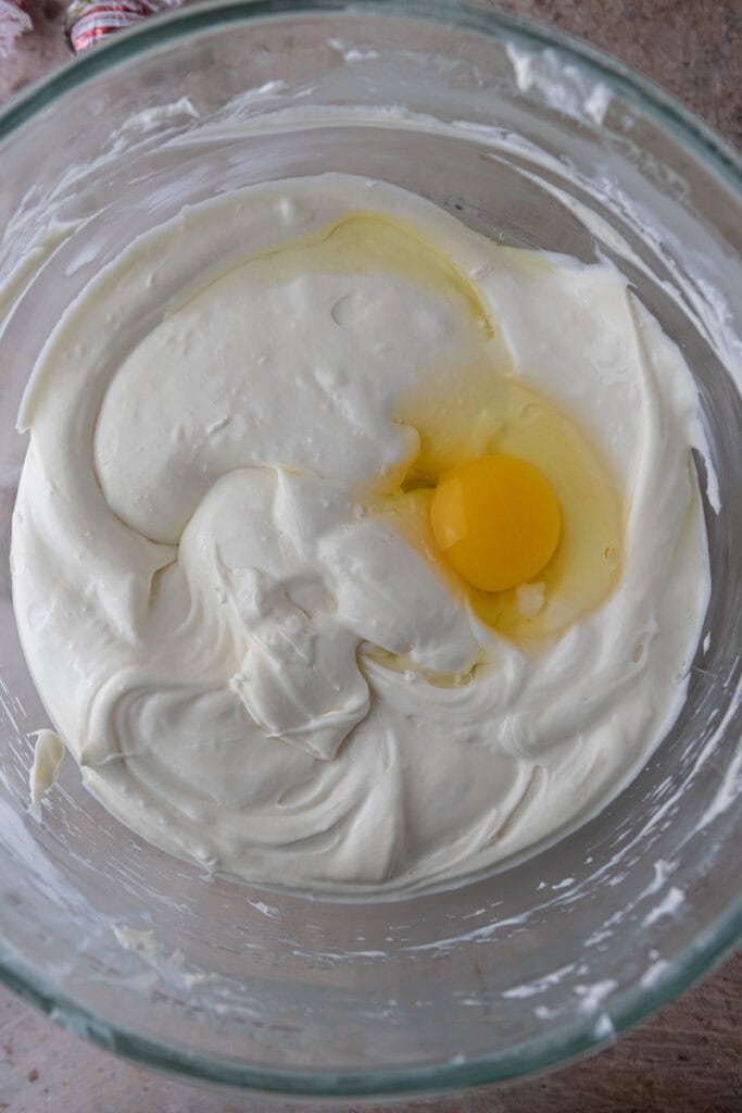 : Creamy cheesecake batter in a bowl with an egg added on top before mixing