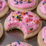 Soft sour cream sugar cookie with pink frosting and rainbow sprinkles, with a bite taken out to show the tender interior.
