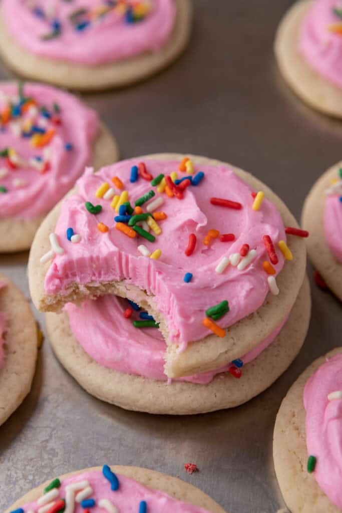 Soft sour cream sugar cookie with pink frosting and rainbow sprinkles, with a bite taken out to show the tender interior.
