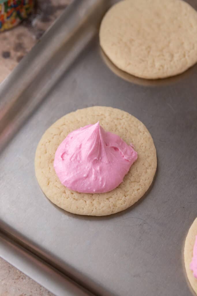 A baked sour cream sugar cookie topped with a spoonful of fluffy pink frosting before spreading.
