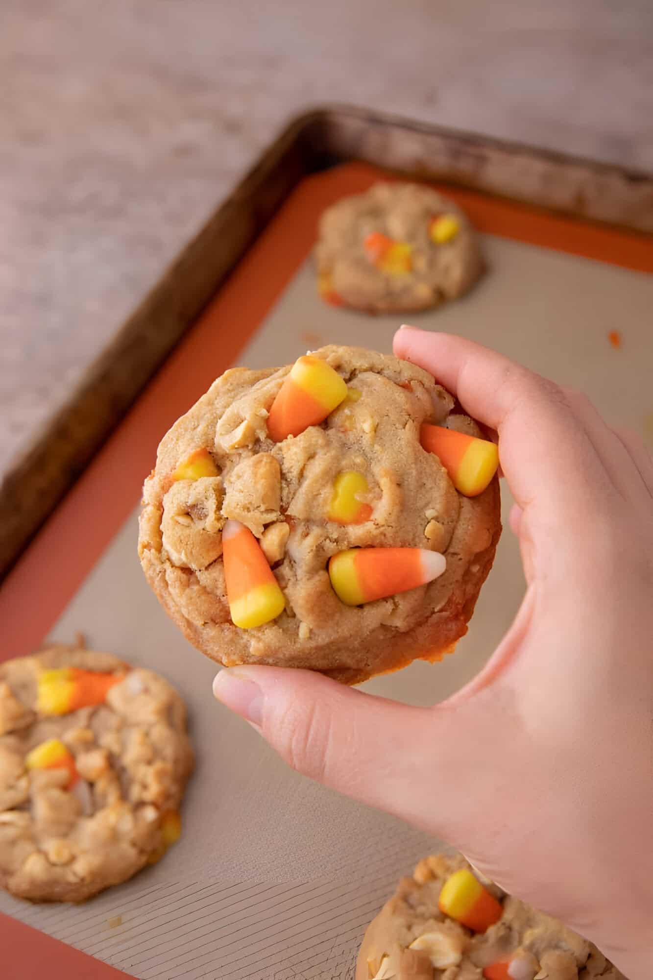 Hand holding a thick candy corn cookie with salted peanuts over a lined baking sheet.