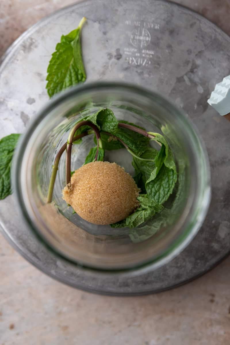 Fresh mint leaves and a scoop of brown sugar in the bottom of a mason jar, seen from above.