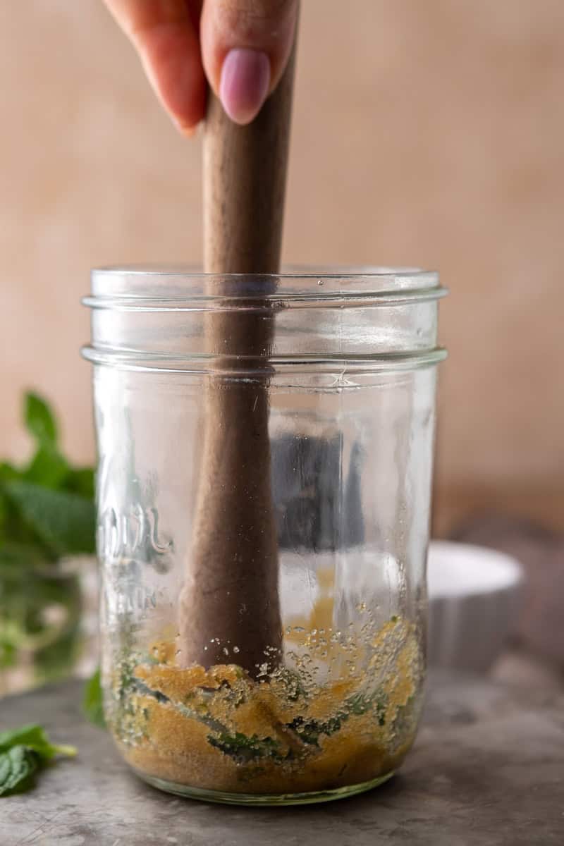 A wooden muddler crushing mint and brown sugar in a mason jar.