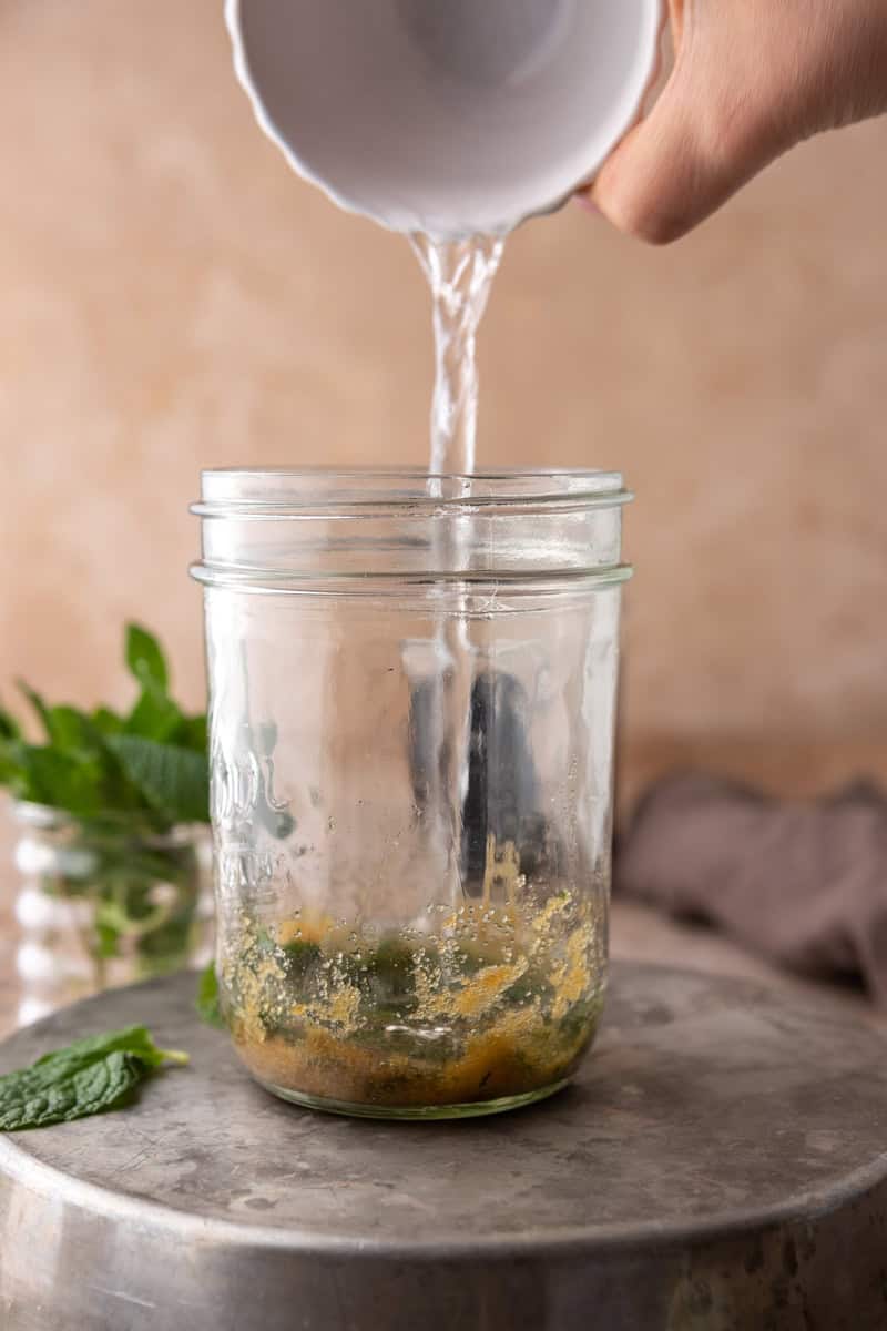 Hot water poured from a small white bowl over the muddled mint and brown sugar in a mason jar.