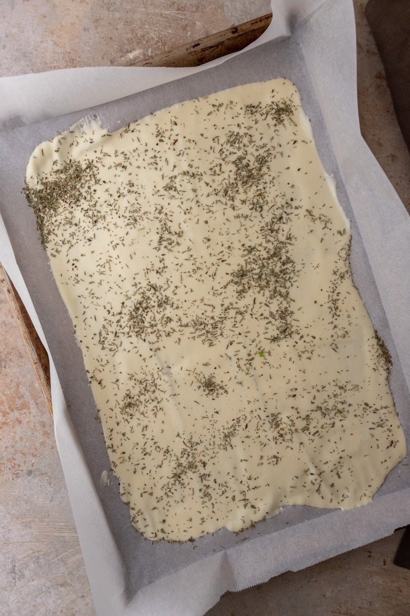 Cracker batter spread thin on a parchment-lined baking sheet and sprinkled with dried herbs.