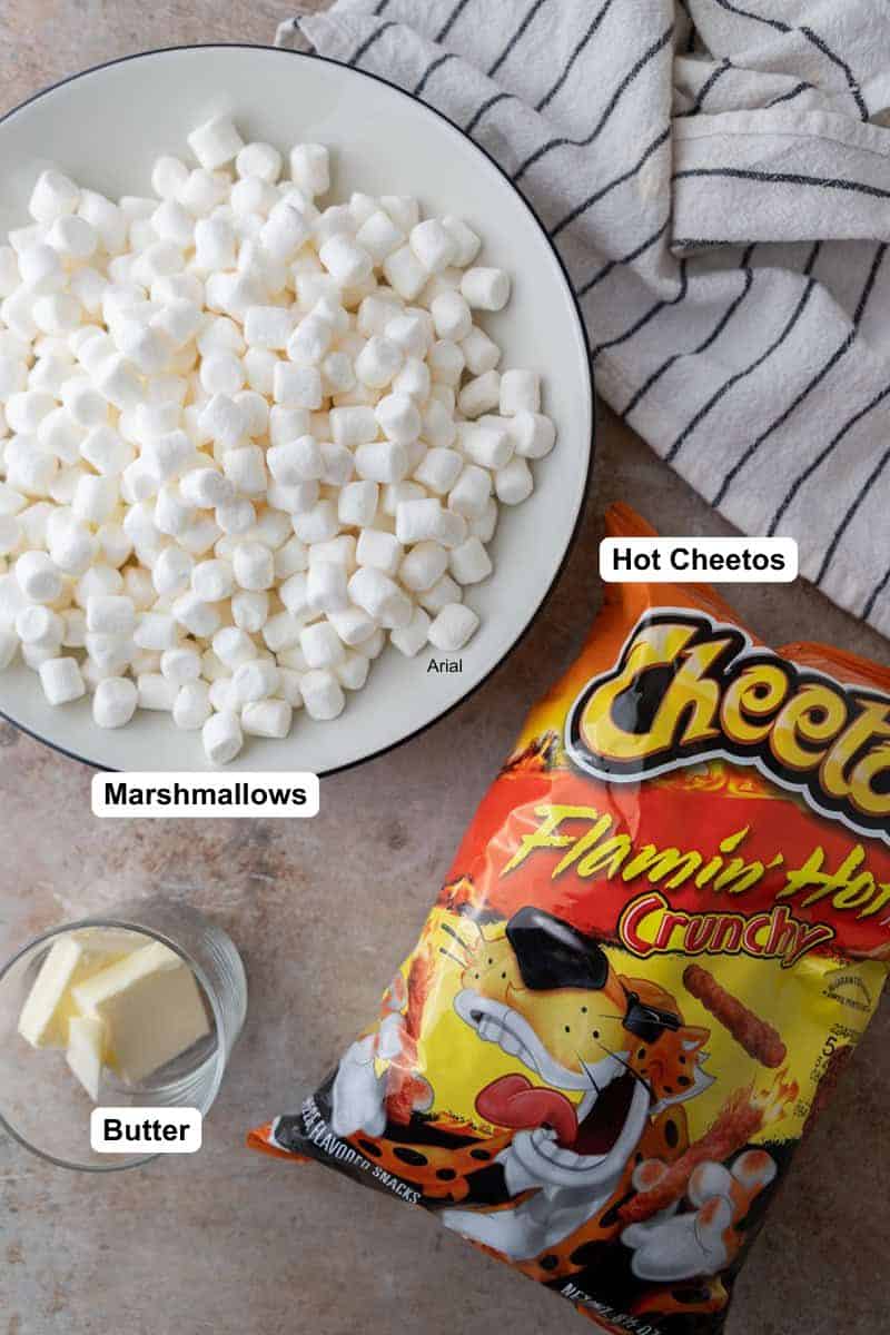 Mini marshmallows in a white bowl, a small dish of butter and a bag of Flamin' Hot Cheetos on a gray countertop.