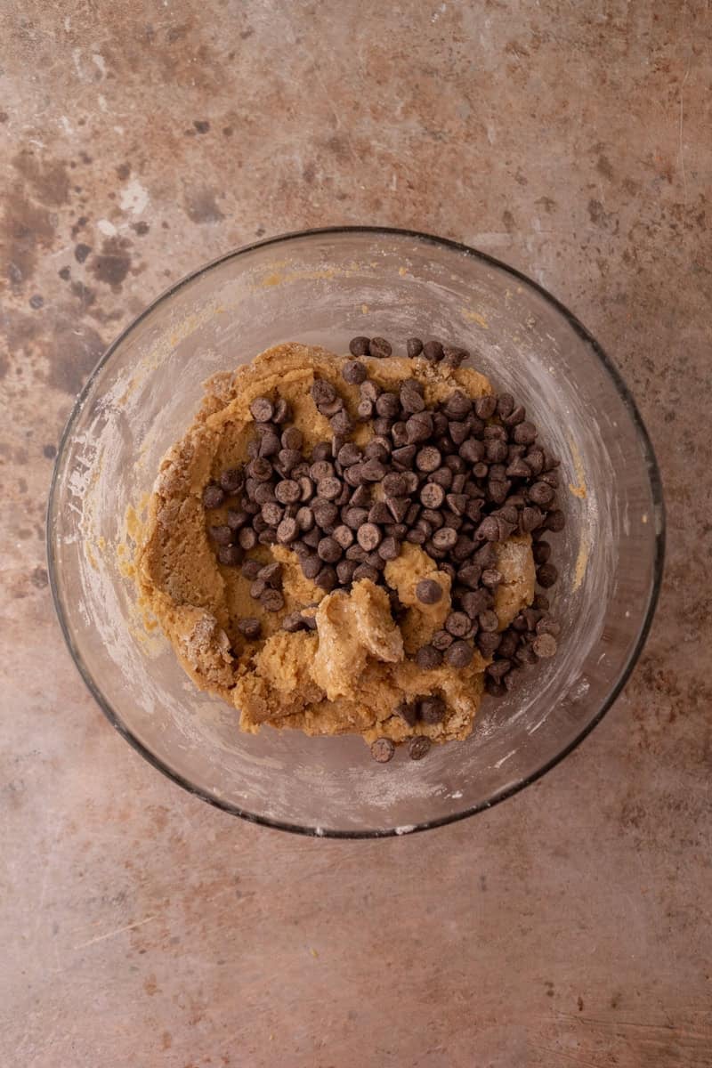 Chocolate chips and dry ingredients added to the peanut butter cookie dough in a glass bowl.