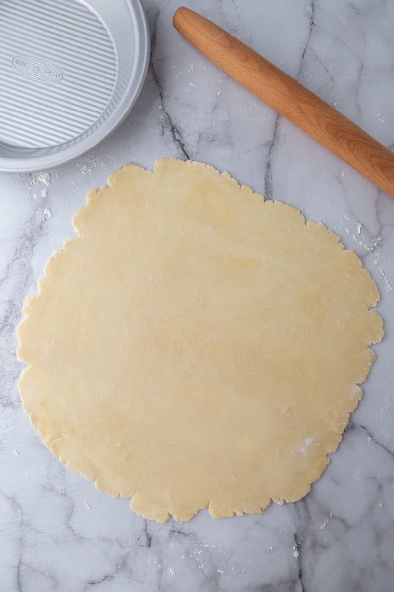 Pie dough rolled out into a large circle on a marble counter next to a rolling pin and pie plate.