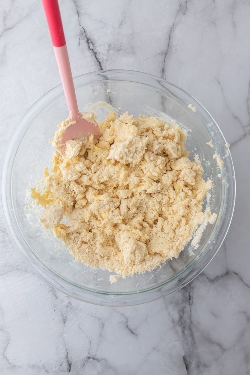 Pink spatula mixing the pie dough in a glass bowl until it starts to clump together.